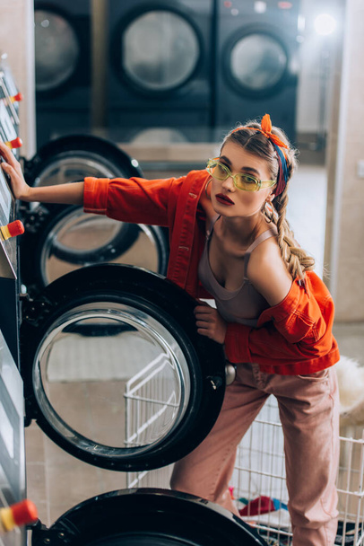 femme élégante dans les lunettes de soleil et bandeau regardant caméra près des machines à laver dans la laverie automatique - Photo, image