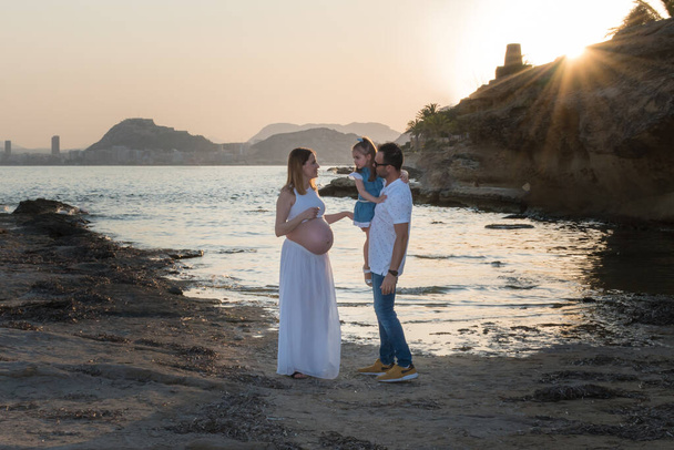 Famiglia incinta con la loro giovane figlia che cammina felicemente sulla spiaggia al tramonto - Foto, immagini