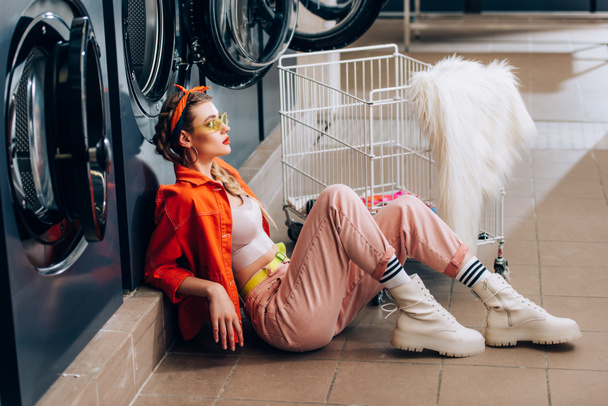 vista lateral de mujer en gafas de sol sentada en el suelo cerca de carro metálico y lavadoras en lavandería - Foto, Imagen