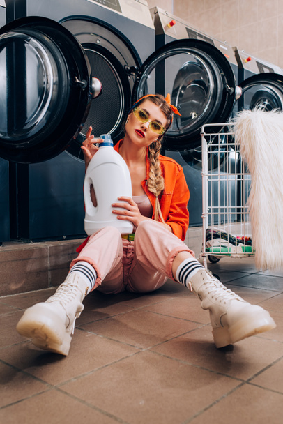 giovane donna in occhiali da sole bottiglia con detersivo e seduta sul pavimento vicino alle lavatrici e carrello in lavanderia - Foto, immagini