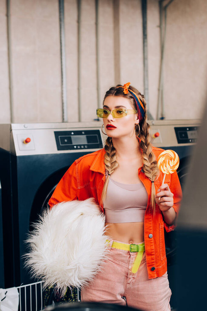 young stylish woman holding lollipop and faux fur coat near washing machines in laundromat - Photo, Image