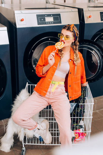 stylish woman in sunglasses biting lollipop near cart with clothing and washing machines in laundromat - Photo, Image