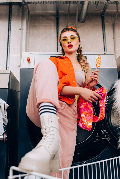 trendy young woman in sunglasses holding lollipop and clothing in laundromat with blurred foreground - Photo, Image