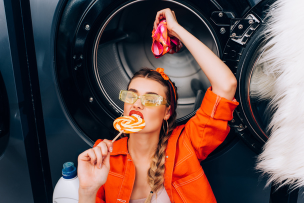 fashionable woman biting sweet lollipop and holding clothing in laundromat - Photo, Image