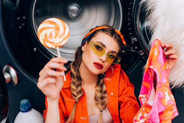 young fashionable woman holding sweet lollipop and clothing in laundromat - Foto, Imagen