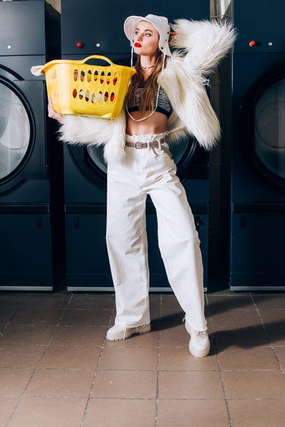 young woman in faux fur jacket and hat holding basket with laundry near washing machines in laundromat - Foto, immagini