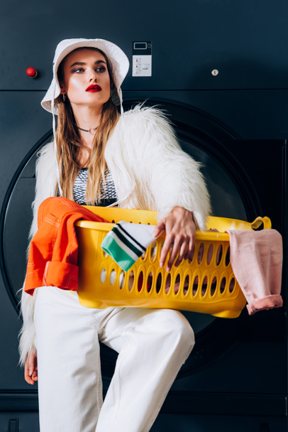 jovem na moda em casaco de pele falso e chapéu segurando cesta com lavanderia perto da máquina de lavar roupa na lavanderia - Foto, Imagem