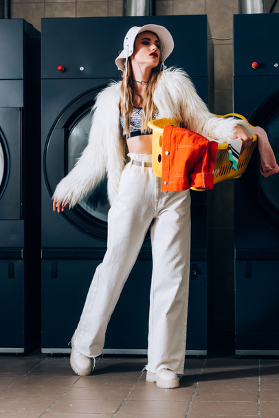 stylish woman in faux fur jacket and hat holding basket with laundry near washing machines in laundromat - Fotó, kép