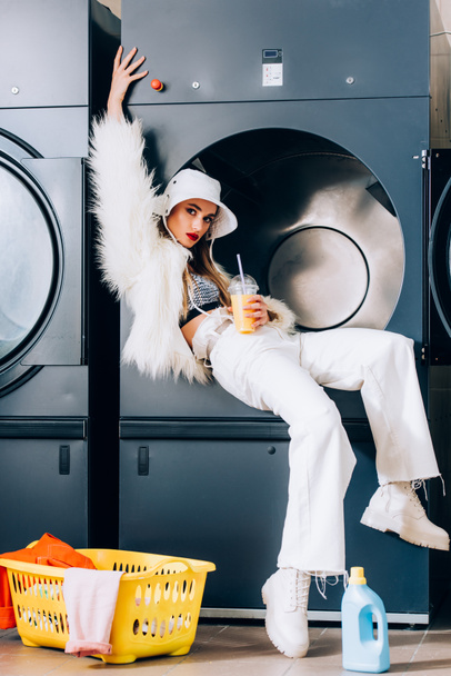trendy woman in faux fur jacket and hat holding plastic cup with orange juice near basket with laundry and washing machine in laundromat - Photo, Image