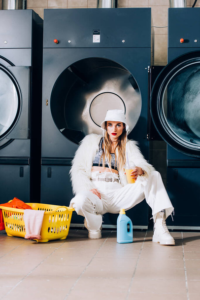 stylish woman in faux fur jacket and hat holding plastic cup with orange juice near basket with laundry and washing machines in laundromat - Photo, Image