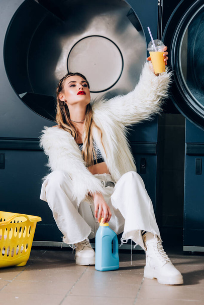 stylish young woman in faux fur jacket holding plastic cup with orange juice near basket with laundry, detergent bottle and washing machines - Photo, image