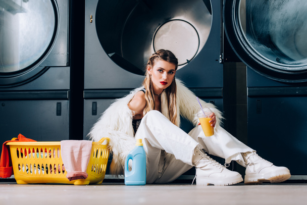 mulher elegante em casaco de pele falso segurando copo de plástico com suco de laranja perto da cesta com lavanderia, garrafa de detergente e máquinas de lavar roupa na lavanderia - Foto, Imagem