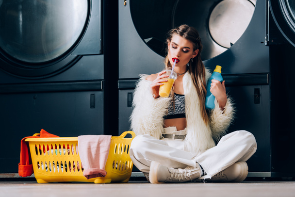 stylish woman in faux fur jacket holding plastic cup with orange juice near basket with laundry, detergent bottle and washing machines in laundromat - Фото, изображение