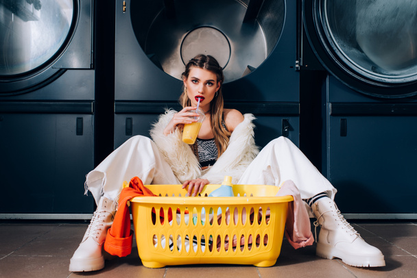 woman in faux fur jacket drinking orange juice and sitting near basket with laundry, detergent bottle and washing machines in laundromat - Photo, Image