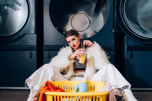 mujer joven sosteniendo una taza de plástico con jugo de naranja y sentada cerca de la canasta con lavandería, botella de detergente y lavadoras en la lavandería - Foto, Imagen