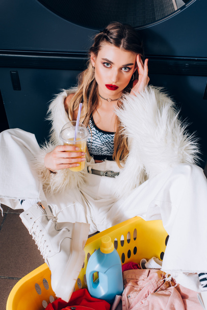 young woman in faux fur jacket sitting with orange juice near basket with dirty clothing  - Photo, Image