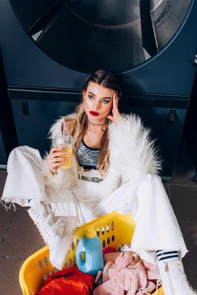 mujer joven con chaqueta de piel sintética sentada con jugo de naranja cerca de la cesta con ropa sucia en la lavandería - Foto, imagen