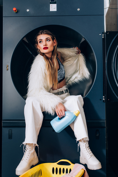 young woman in white faux fur jacket sitting in washing machine and holding bottle with detergent in laundromat - Fotó, kép