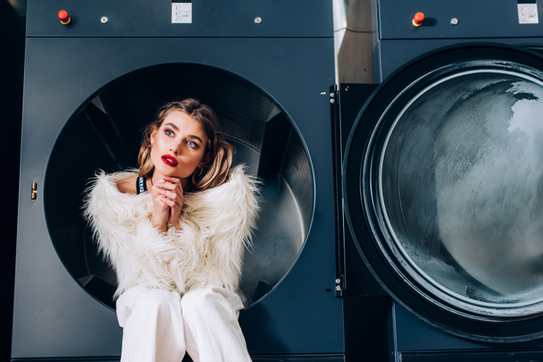 mujer de ensueño en chaqueta de piel sintética blanca sentada en una lavadora pública  - Foto, Imagen
