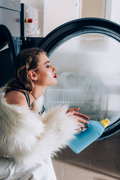Seitenansicht einer jungen stilvollen Frau, die eine Flasche mit Waschmittel in der Hand hält und in einer öffentlichen Waschmaschine sitzt  - Foto, Bild