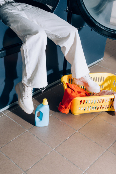 cropped view of woman in white pants sitting in public washing machine near basket with laundry and detergent bottle - Photo, Image