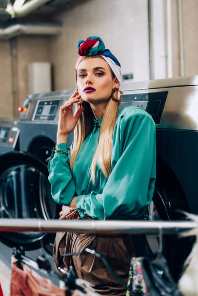 stylish woman in turban standing near washing machines in public laundromat  - Zdjęcie, obraz