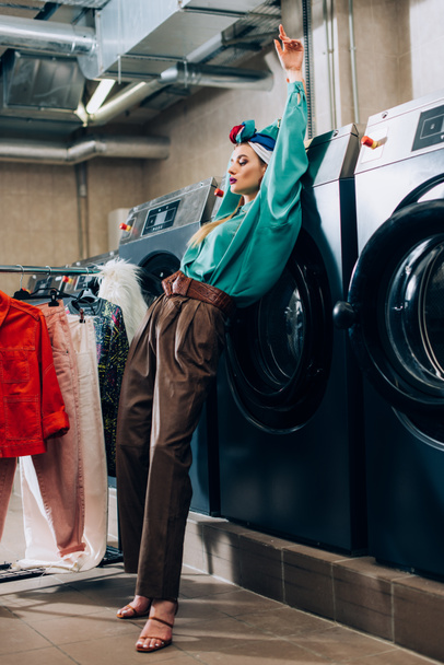 trendy woman in turban standing near clothing rack and washing machines in laundromat  - Foto, imagen