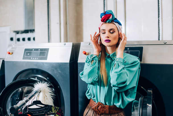 femme élégante touchant turban et debout près des machines à laver dans la laverie automatique moderne  - Photo, image