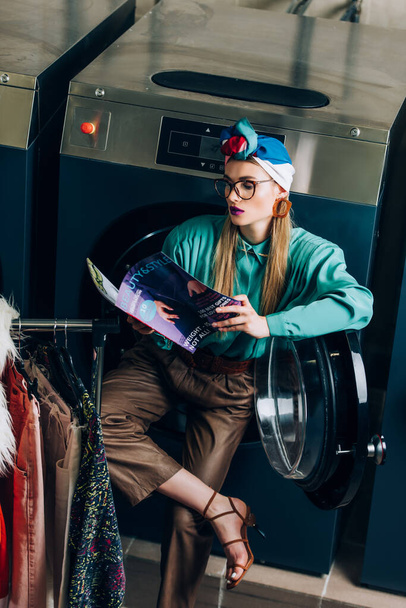 young and stylish woman in turban and glasses reading magazine in laundromat  - Valokuva, kuva