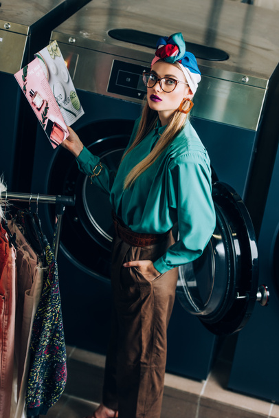 mujer joven y elegante en turbante y gafas que sostienen la revista y de pie con la mano en el bolsillo en la lavandería  - Foto, Imagen