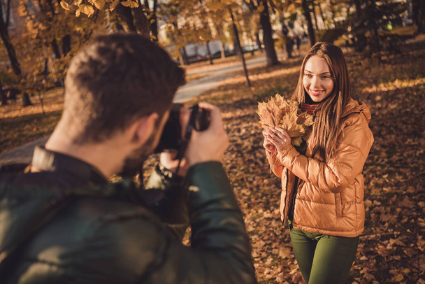 Photo of young guy taking photo digital camera his beautiful girl hold maple leaves in fall september town park - 写真・画像