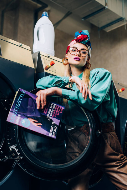 low angle view of young woman in glasses and turban holding magazine near washing machines in public laundromat  - Fotografie, Obrázek