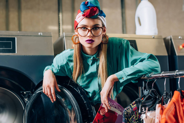 woman in glasses and turban holding magazine near modern washing machine in public laundromat  - Fotografie, Obrázek