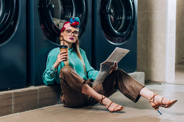 trendy woman in glasses and turban holding paper cup and newspaper while sitting in laundromat  - Foto, afbeelding