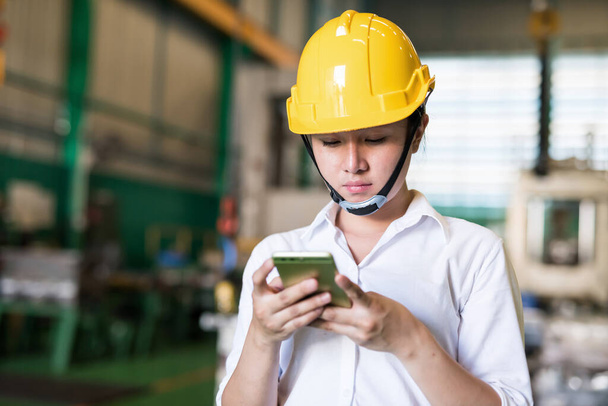 Factory engineer with hardhat check corporate app and chat on smartphone in factory warehouse. Manufacturing industry and technology. Industry with inventory concept. - Φωτογραφία, εικόνα