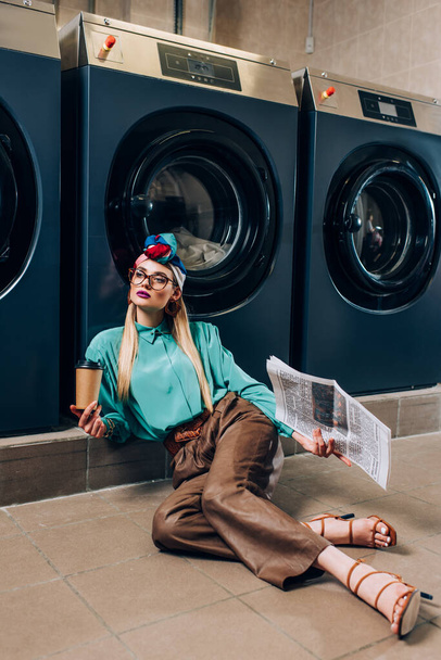 jeune femme dans des lunettes et turban tenant tasse en papier et journal tout en étant assis sur le sol dans la laverie automatique  - Photo, image