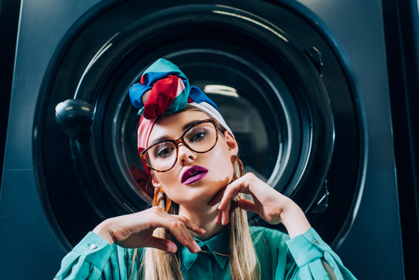 stylish young woman in glasses and turban looking at camera near washing machine - Zdjęcie, obraz