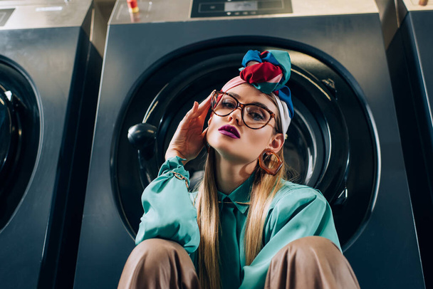 mujer joven de moda en gafas y turbante mirando a la cámara cerca de las lavadoras - Foto, Imagen