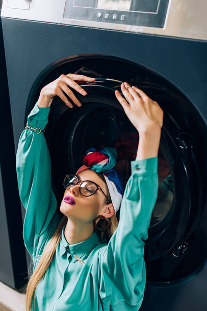 stylish young woman in glasses and turban touching door of washing machine in laundromat - Φωτογραφία, εικόνα
