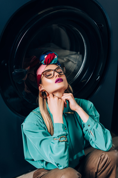élégant jeune femme dans des lunettes et turban assis avec les yeux fermés près de la machine à laver - Photo, image