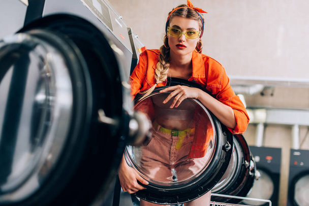 stylish woman in headband and sunglasses near washing machines in modern laundromat with blurred foreground - Фото, изображение