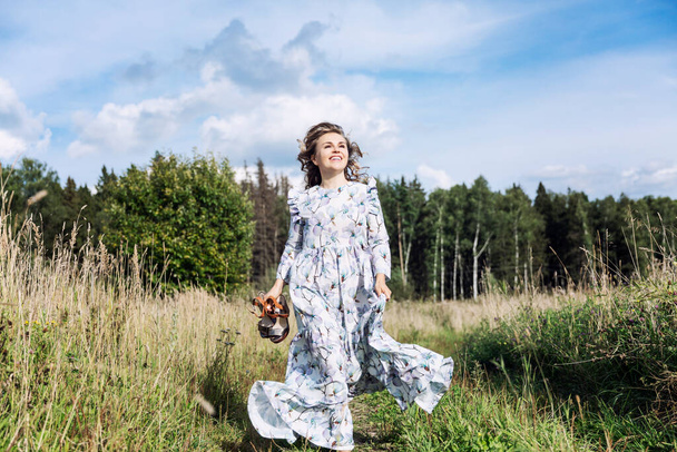 Una giovane donna ridente con un lungo abito romantico corre sul campo in una giornata di sole. La felicità è in unità con la natura. - Foto, immagini