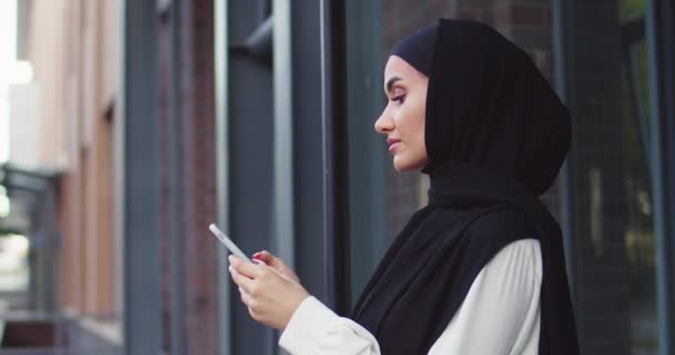 Close up portrait of Arabic woman in traditional headscarves looking at phone and smiling. Pretty muslim female in hijabs reading messages, texting, browsing Internet on smartphone after shopping. - Filmagem, Vídeo
