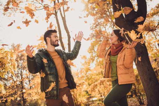 Photo of positive cheerful couple girl guy throw september yellow leaves in fall october outdoors city forest wear coats - Foto, afbeelding
