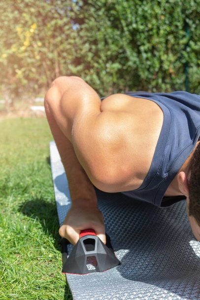 detail of the muscular arm of a boy with sports clothes doing crosstraining and push-ups with muscle clamps in the garden of his house in a sunny day - Photo, Image