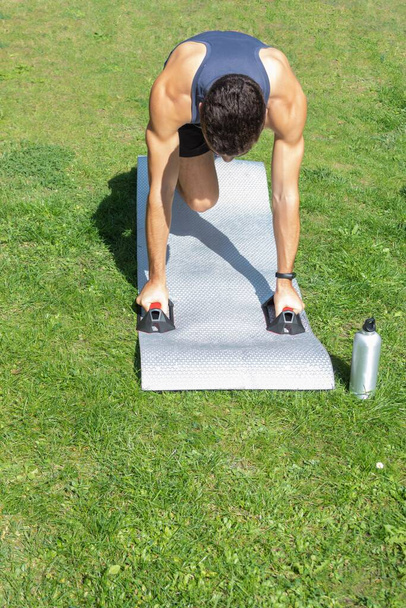 jongen in sportkleding doet push-ups met crosstraining grepen push up bars in zijn achtertuin waar u een fles water op een zonnige dag kunt zien - Foto, afbeelding