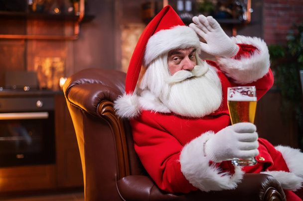 Santa Claus drinks beer while sitting in a leather armchair - Fotó, kép