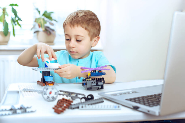 Cheerful smart preschool boy sitting at the table and constructing a robotic device at home. Online education, E-learning concept, distance communication with laptop - Photo, Image