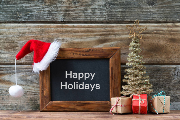 Vrolijke kerstversiering, krijtbord met tekst Vrolijke feestdagen en een rode kerstmuts, kleine kerstboom en geschenken op een houten achtergrond - Foto, afbeelding