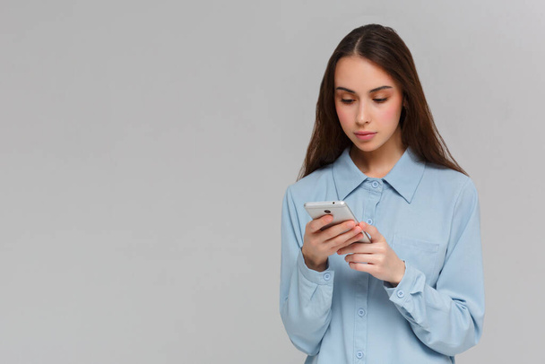 Content teenager with long hair, holds modern cell phone, scrolls through social networks, has cheerful expression, isolated over gray background, recieves message - Valokuva, kuva